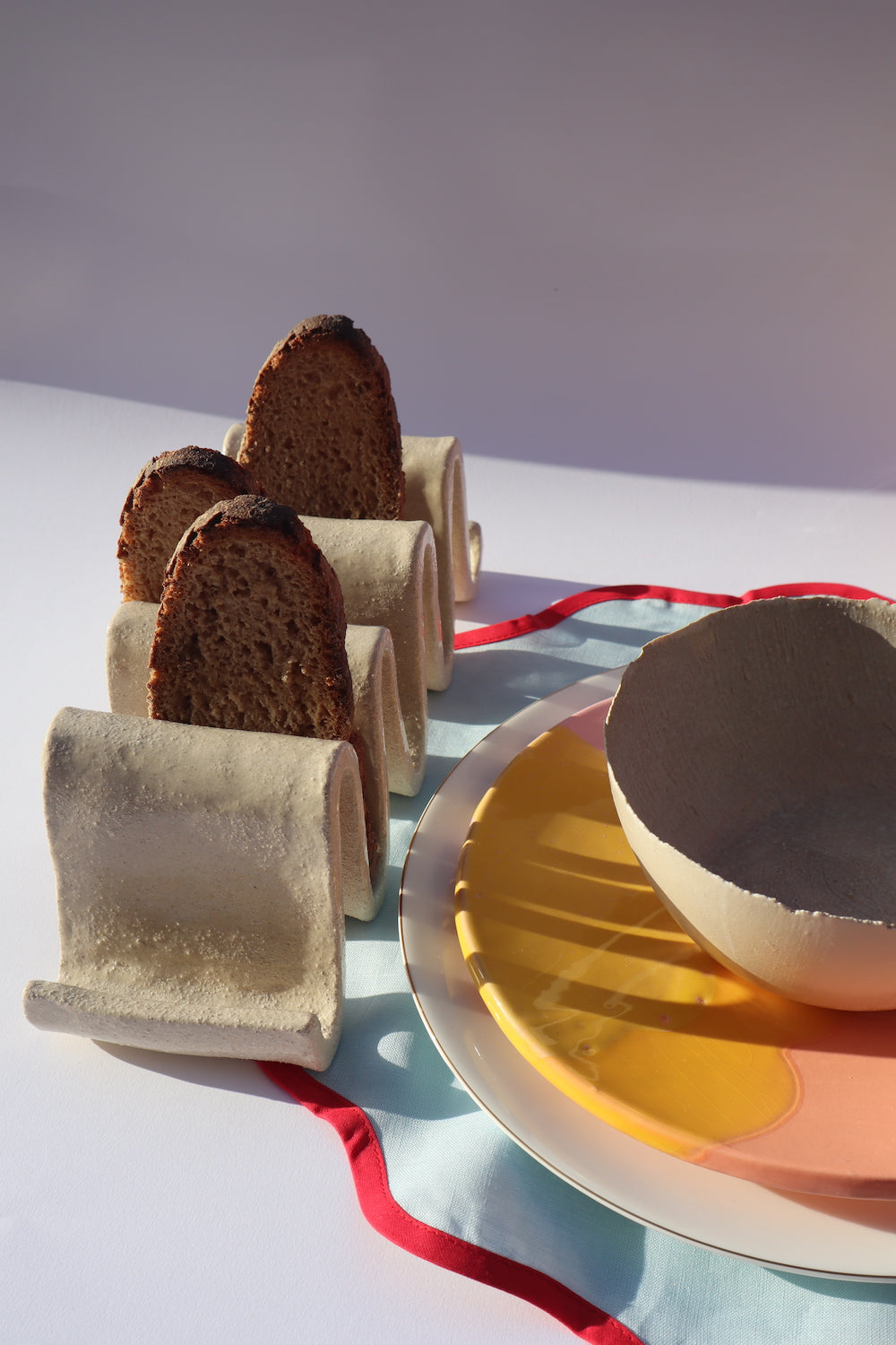 Table de petit-déjeuner ensoleillé. Porte-pain en grès blanc chamotté, en forme de vagues. Fait à la main à Paris par l'un de nos artisans. Doté de plusieurs usages, il pourrait tout à fait servir de porte-pain pour y déposer ses tartines grillées le matin, de porte livres sur un bureau ou une table de chevet, ou pour trier votre courrier dans votre entrée.